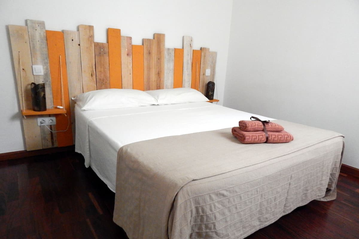 Een net opgemaakt bed met een beige sprei en kussens, een houten hoofdbord met een verticaal patroon, en een paar roze handdoeken op het bed.