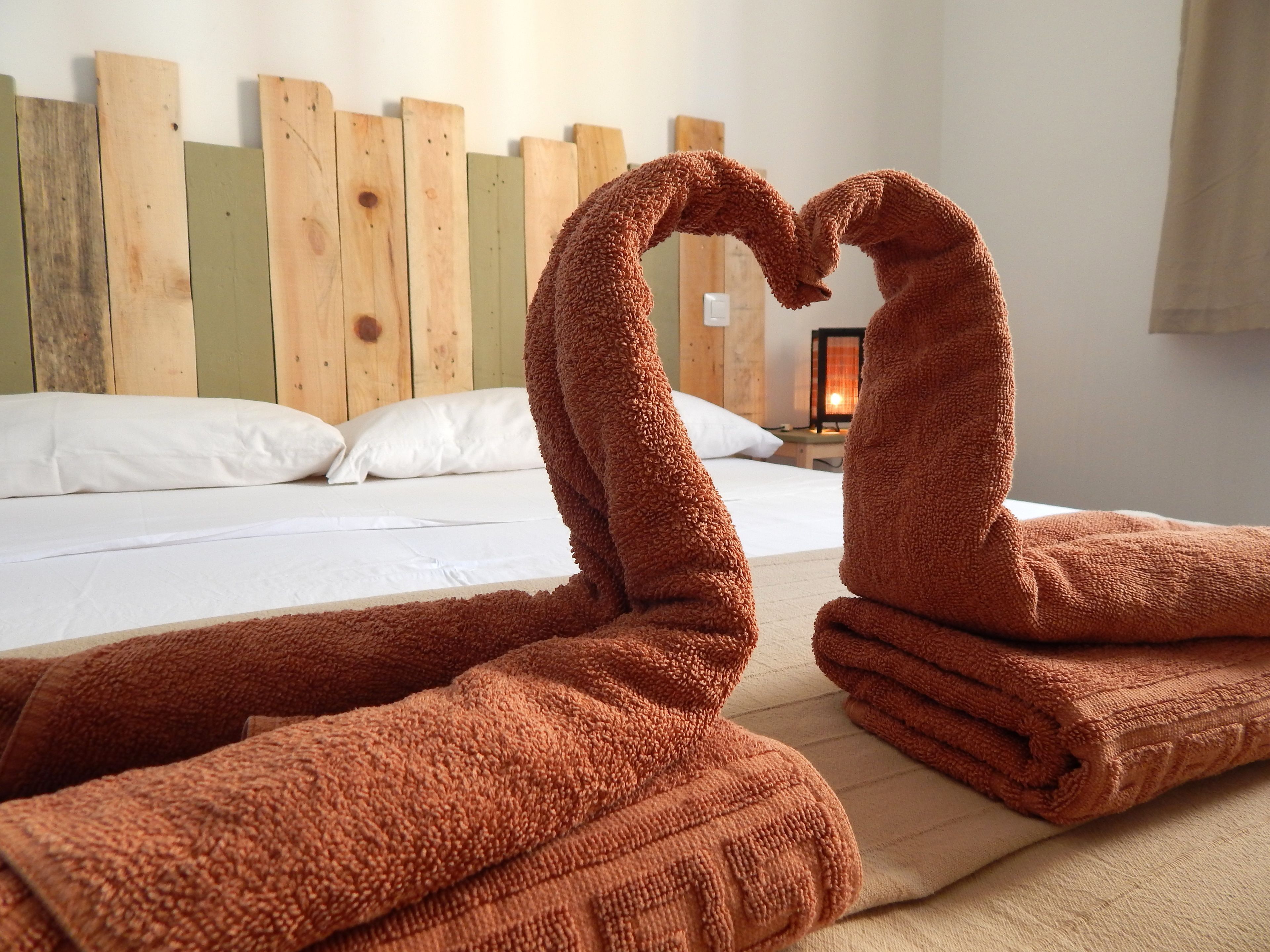 Habitación de hotel con una cama, ropa de cama blanca, cabecera de madera y dos toallas dispuestas en forma de corazón.