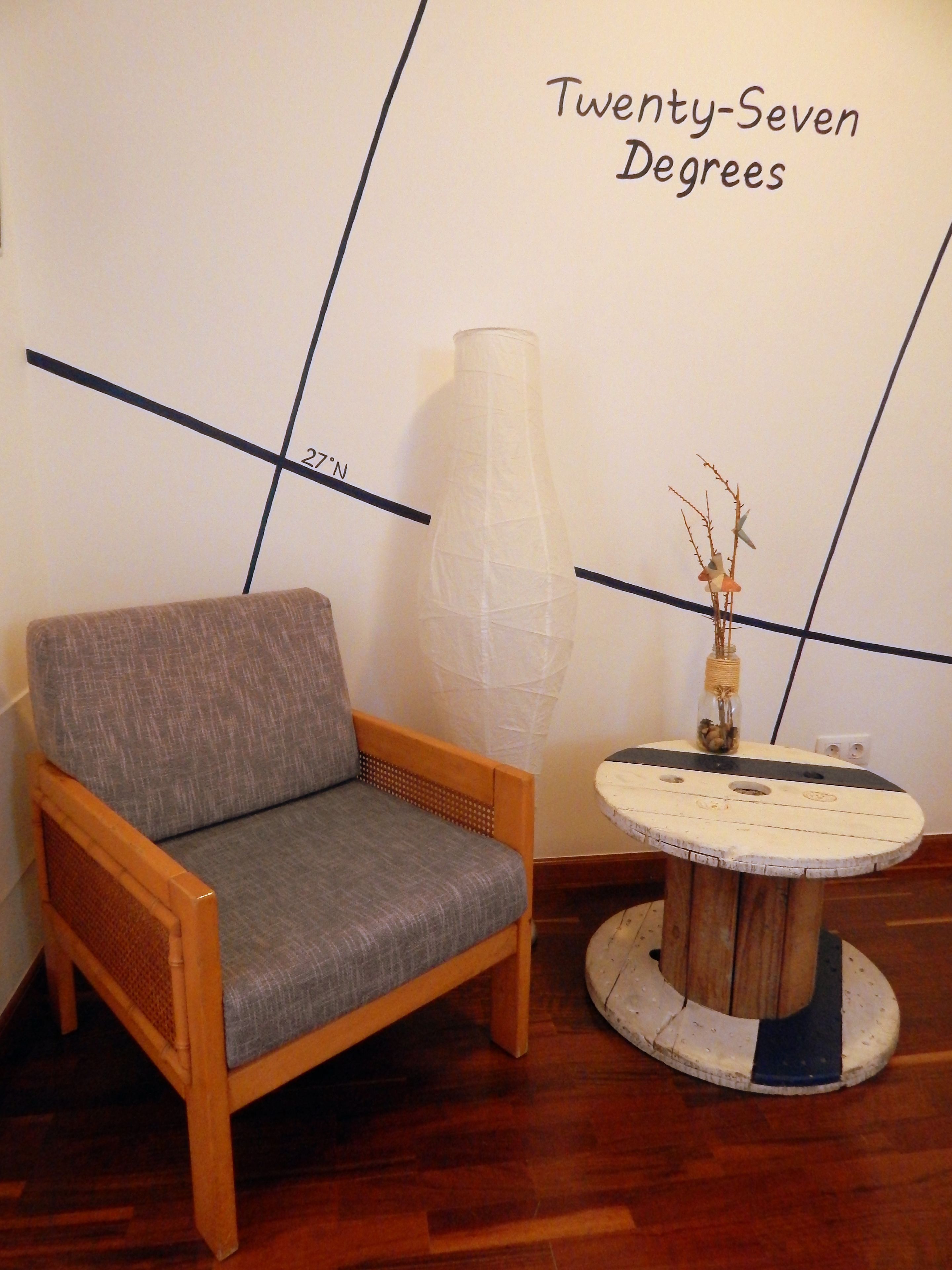 A room with a wooden chair and a small round table with a vase. The wall has a geometric design and text reading "Twenty-Seven Degrees".