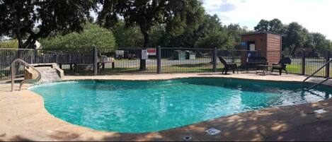 A rectangular pool with a small slide, surrounded by a fence, and shaded by trees.