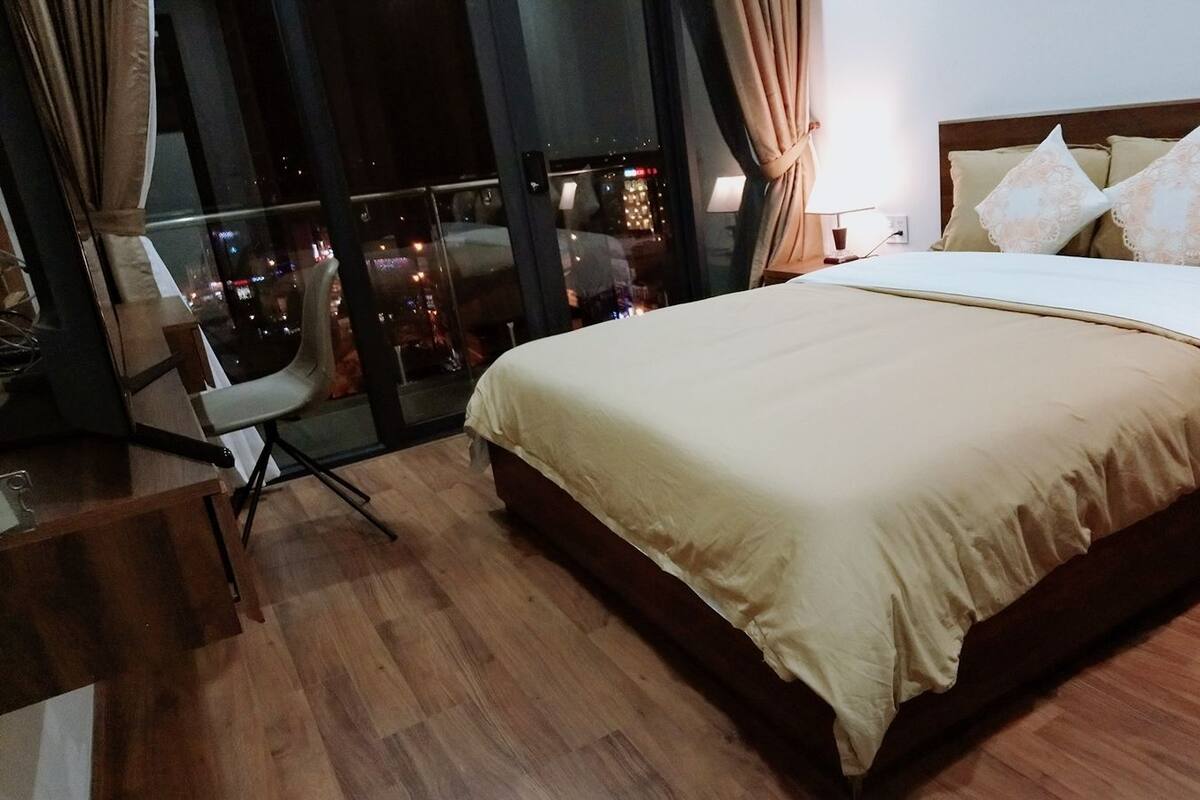 Habitación de hotel con una cama grande, una silla, un escritorio y vistas a la ciudad de noche.