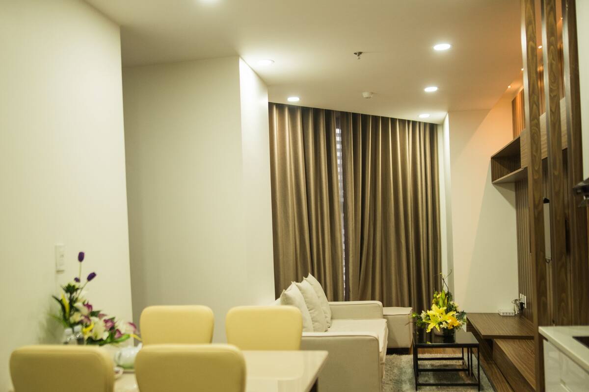 Una sala de estar moderna con sofá, mesa de comedor y sillas. Hay una zona de cocina con armarios de madera y encimera.