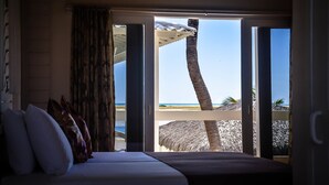 Ein Schlafzimmer mit großem Fenster, das Blick auf einen Strand bietet, ein Bett mit Kissen sowie eine Sicht auf Palmen und ein Reetdachhaus.