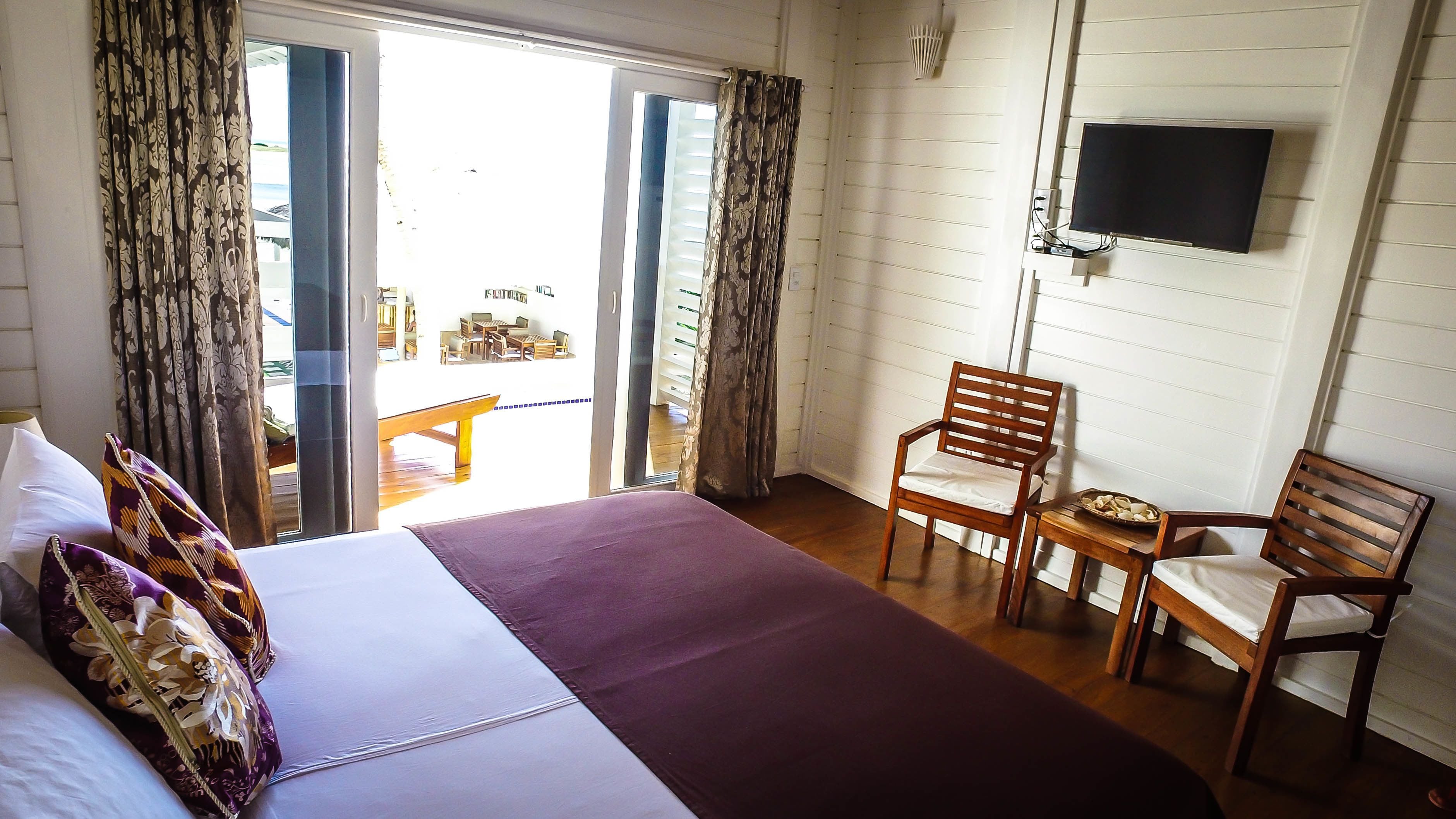 A hotel room with a bed, two wooden chairs, a TV mounted on the wall, and a view of the outside balcony.