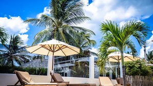Ein Poolbereich mit Liegestühlen und Sonnenschirmen, umgeben von Palmen und blauem Himmel.