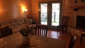 L’intérieur chaleureux d’un chalet avec une cheminée en pierre, des murs en bois, un coin repas doté d’une table en granit et un canapé agrémenté de coussins à motifs.