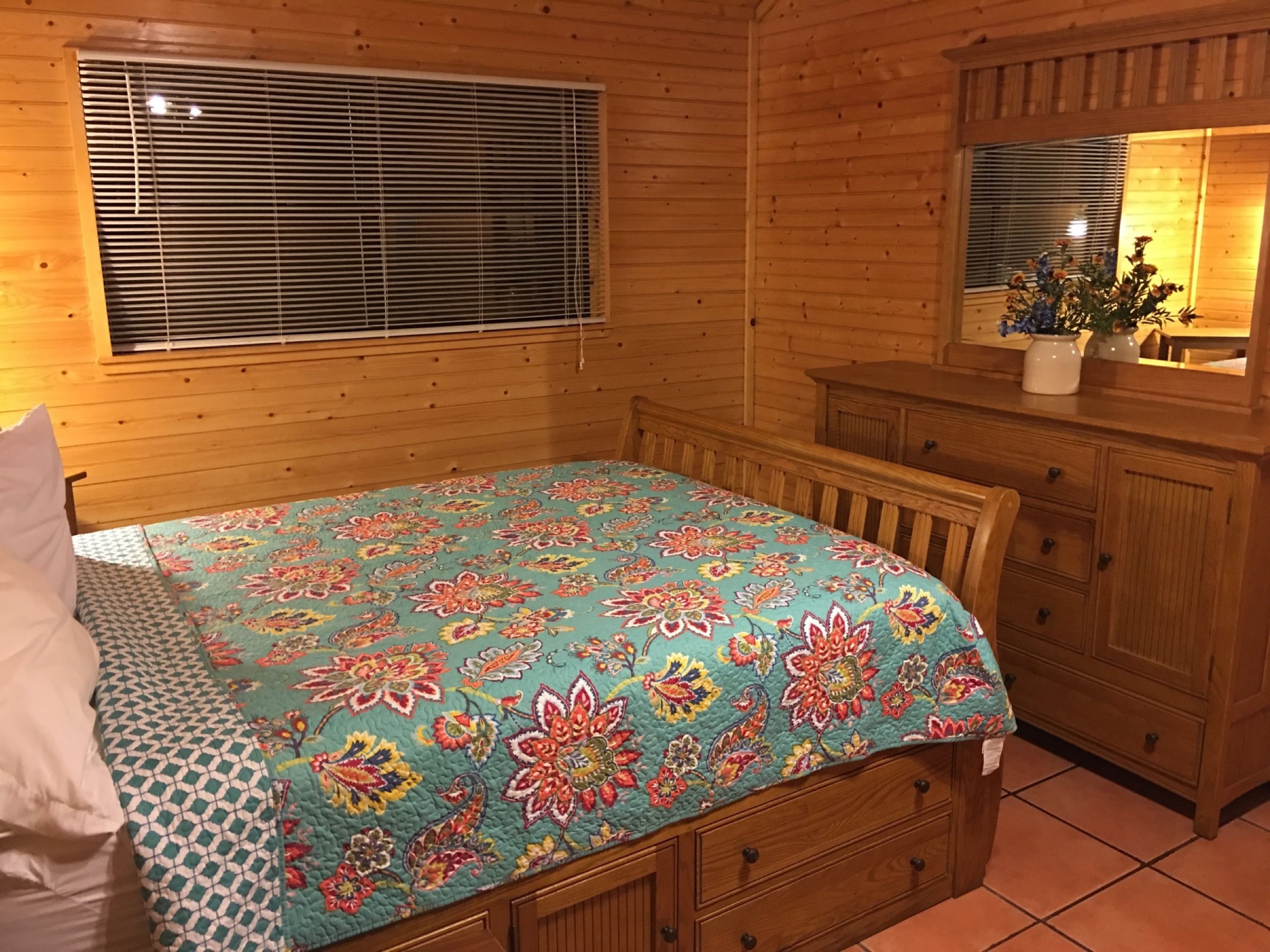 A wooden cabin room with a bed, a dresser, and a window with blinds.
