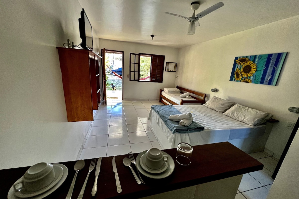 A room with a bed, a dining table set with cutlery, a ceiling fan, and a painting on the wall.