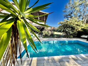 Ein Schwimmbecken mit einer Deckplatte aus Steinplatten, umgeben von Grünpflanzen und einem Gebäude mit Holzbauweise.