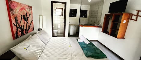 A hotel room with a bed, a painting, a ceiling fan, a bathroom, and a kitchenette.