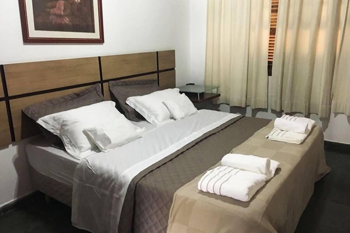Ein Hotelzimmer mit einem Doppelbett, zwei Kissen, einem Nachttisch, einer Lampe und einem mit Vorhängen versehenen Fenster.
