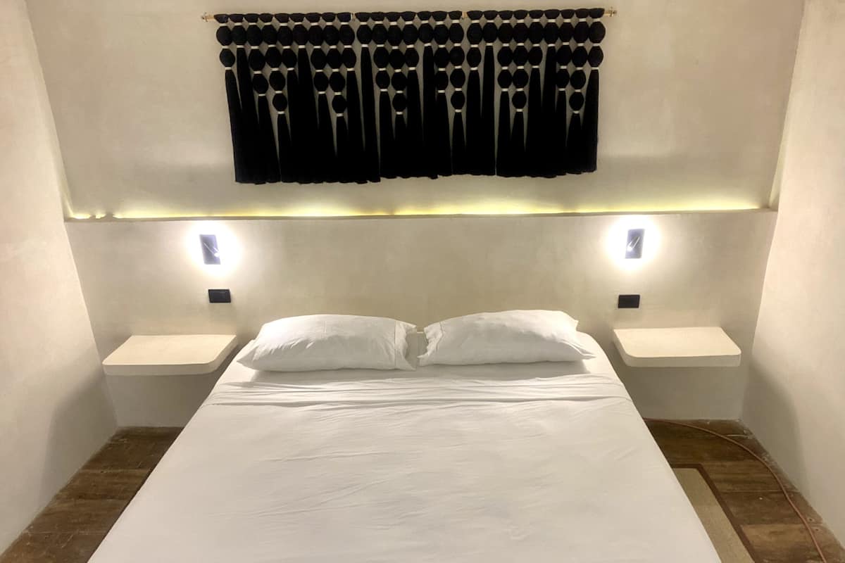 Ein ordentlich bezogenes Bett mit weißen Laken, zwei Nachttische mit Lampen und ein schwarzer Vorhang mit kreisförmigen Mustern.