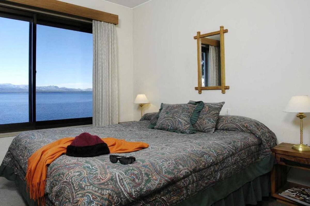 A bedroom with a large window offering a view of a body of water and mountains. There is a bed with patterned bedding, a bedside table with a lamp, and a mirror on the wall.