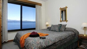 A bedroom with a large window offering a view of a body of water and mountains. There is a bed with patterned bedding, a bedside table with a lamp, and a mirror on the wall.