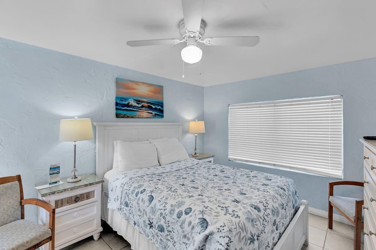 A bedroom with a bed, two bedside tables with lamps, a ceiling fan, a painting, and a window with blinds.