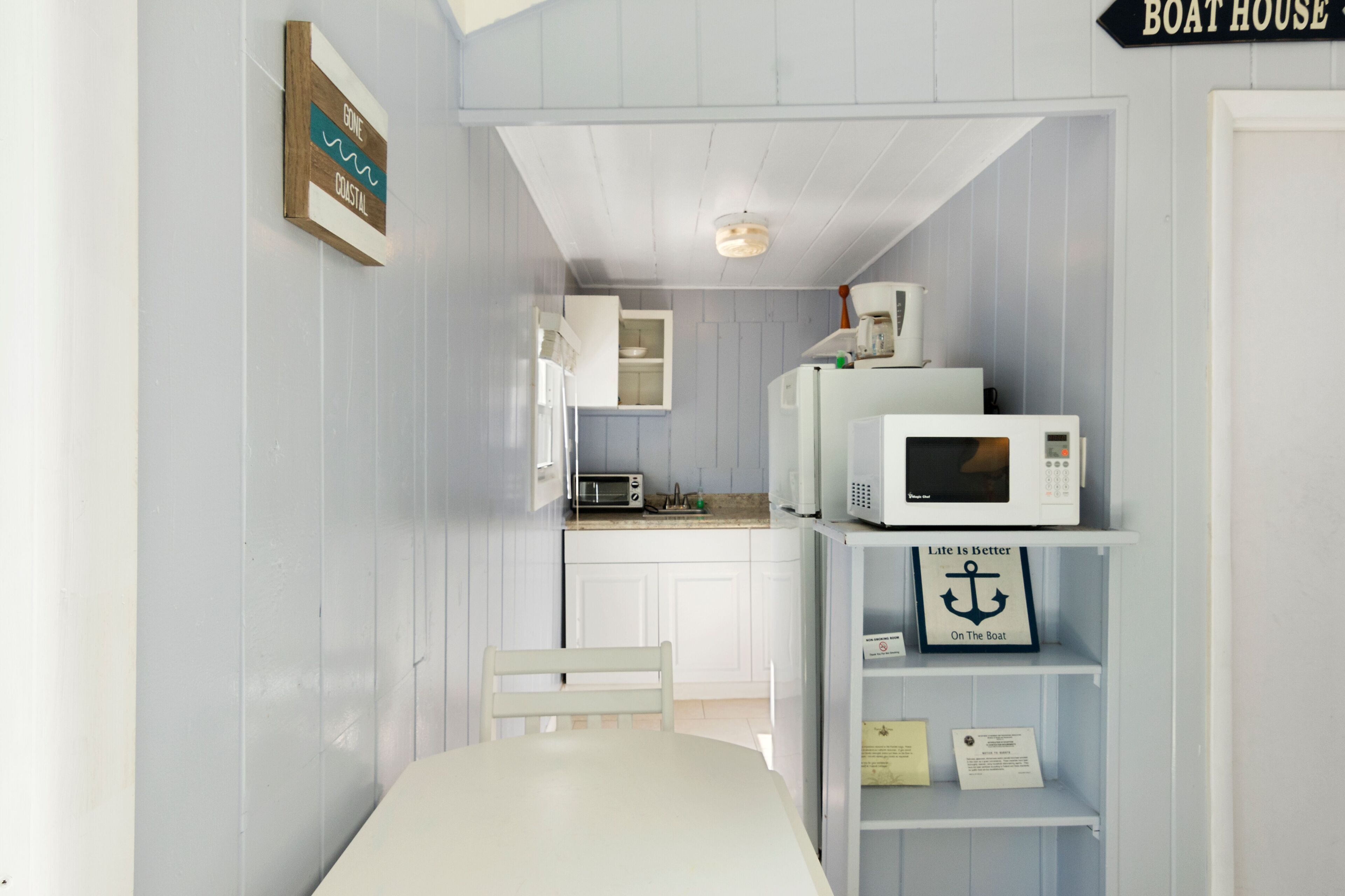 Uma cozinha compacta com armários brancos, micro-ondas e um refrigerador pequeno. Há uma placa na parede com a inscrição 
