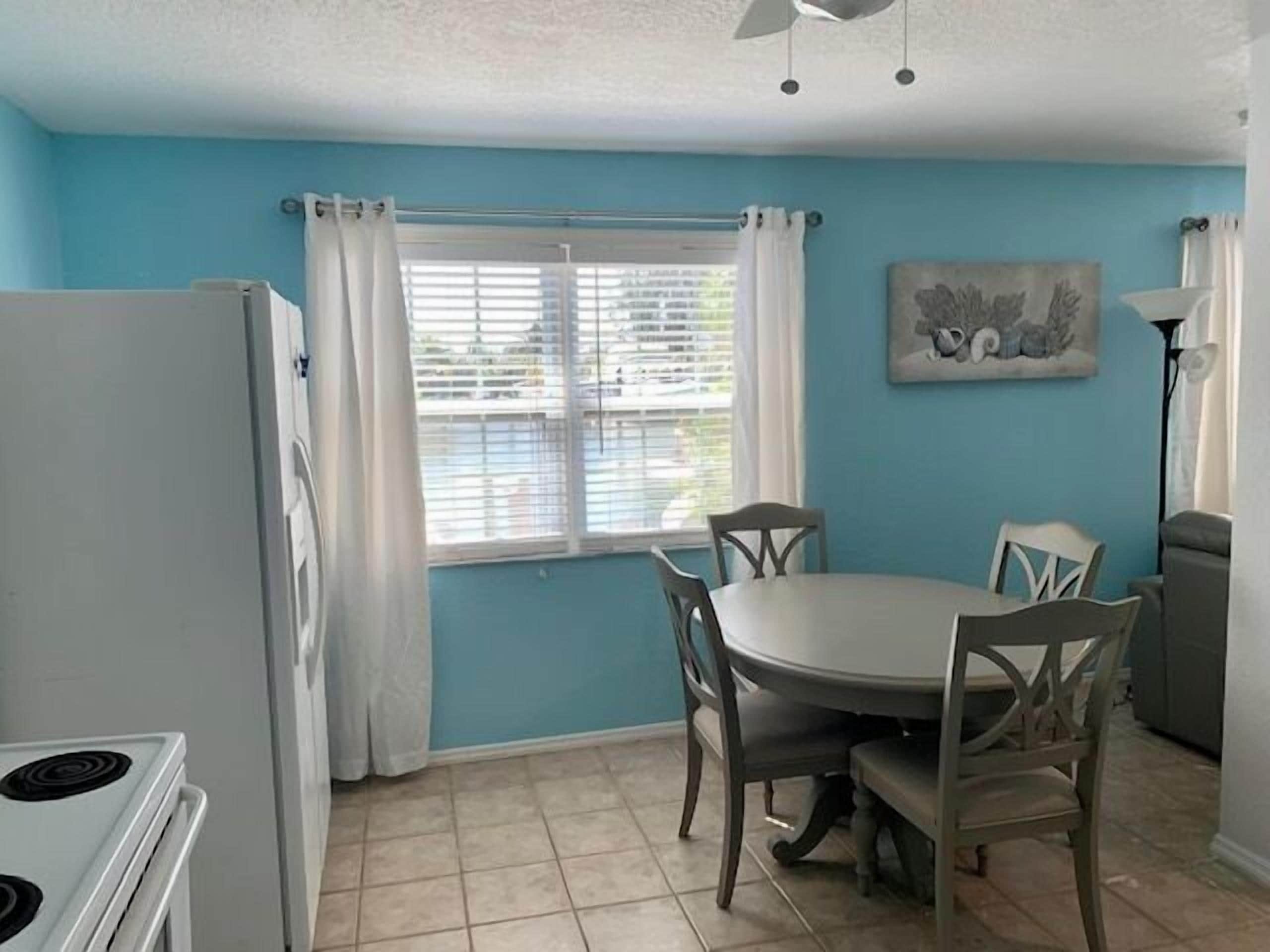 A compact kitchen with a dining area, featuring a white refrigerator, a stove, a dining table with four chairs, and a window with curtains.