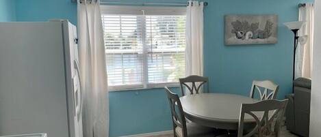 A compact kitchen with a dining area, featuring a white refrigerator, a stove, a dining table with four chairs, and a window with curtains.