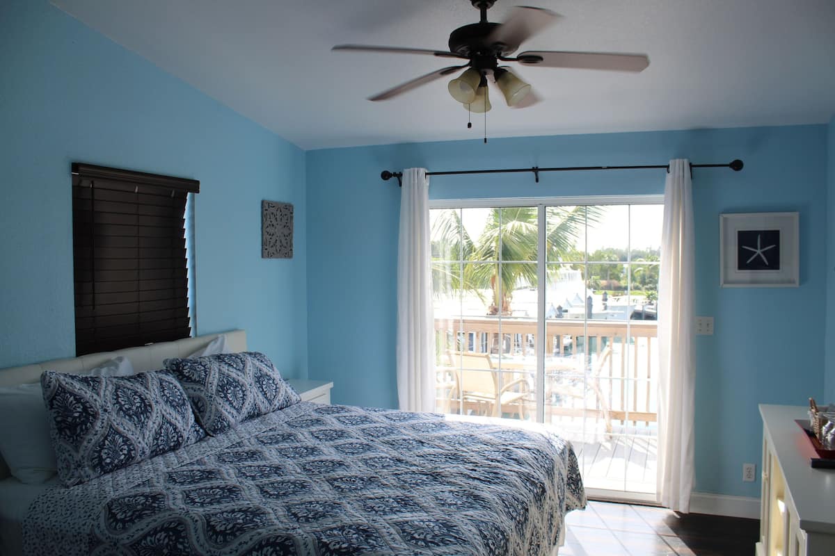 A bedroom with a bed, a ceiling fan, a window with curtains, and a view of the outside.