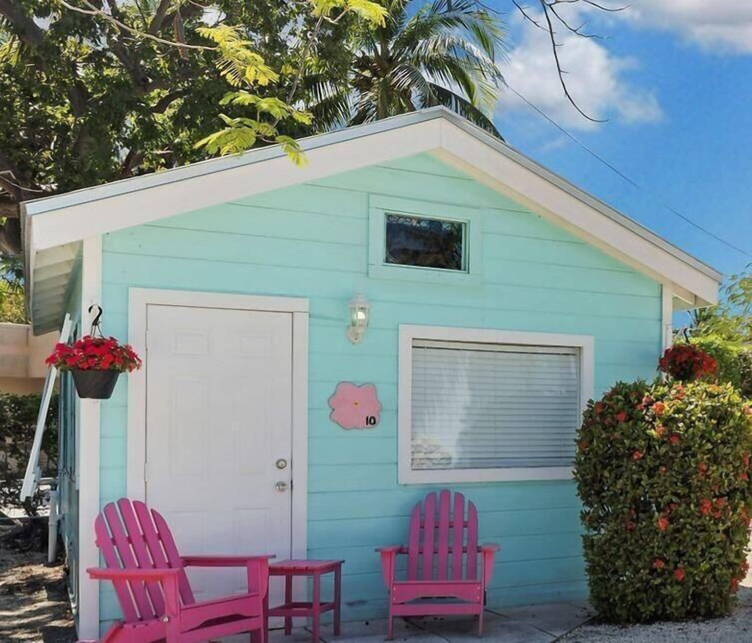 Ein kleiner, hellblauer Schuppen mit weißer Tür und Fenster, zwei pinkfarbene Adirondack-Stühle und ein Blumentopf mit roten Blüten.