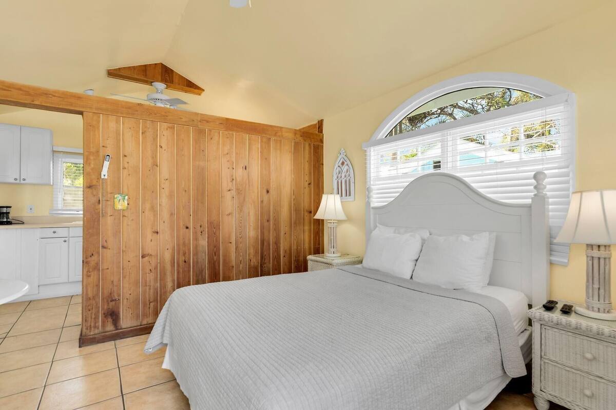 A bedroom with a bed, a nightstand, a lamp, a window with blinds, and a wooden door.