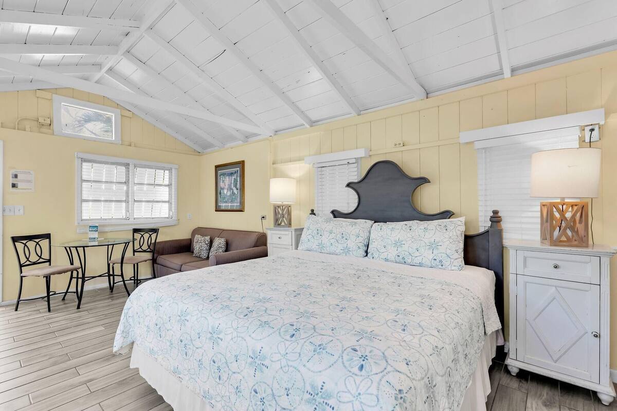 A bedroom with a large bed, a nightstand, a dresser, a window with blinds, a chair, and a small table.