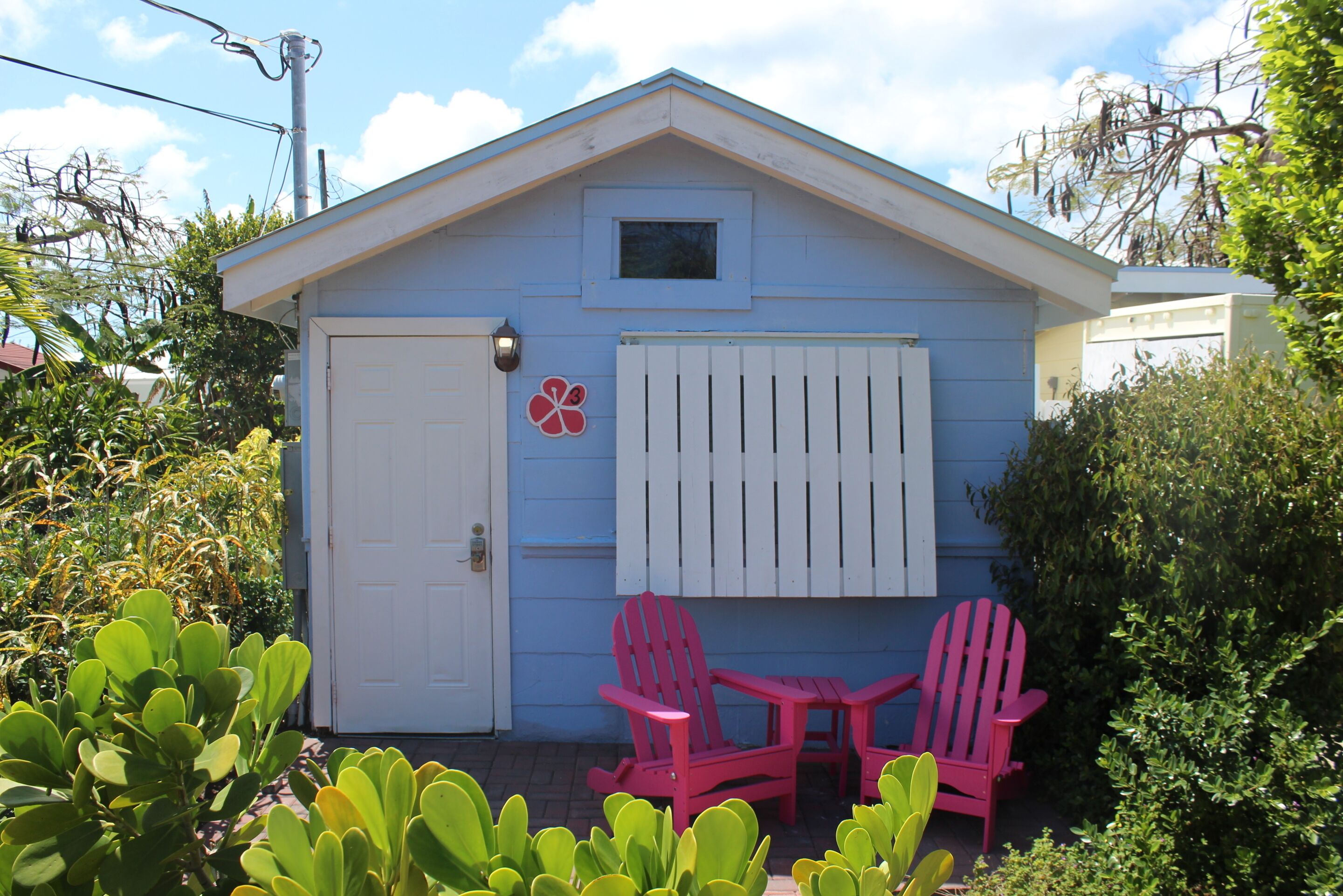 A small blue house with a white door and window, two pink Adirondack chairs, and a small garden area.