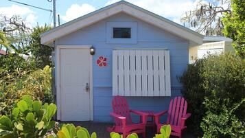 A small blue house with a white door and window, two pink Adirondack chairs, and a small garden area.