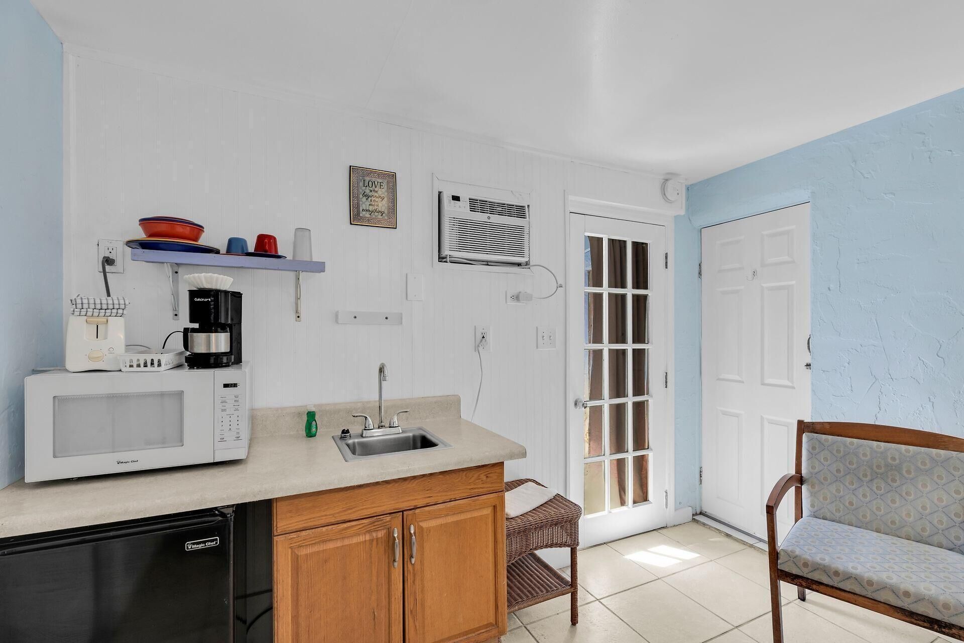 A compact kitchen with a microwave, coffee maker, and sink. There's a wicker chair and a door leading outside.