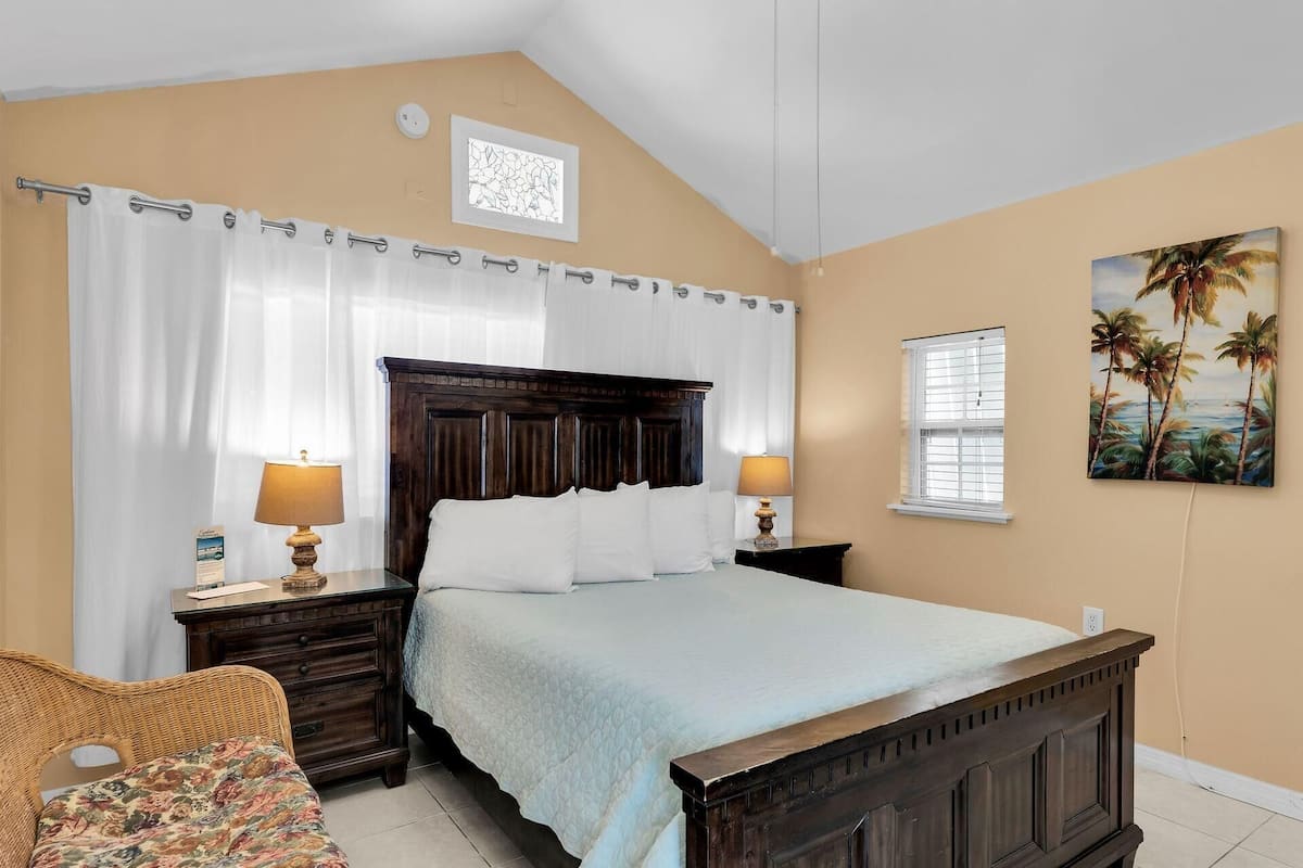 A bedroom with a wooden bed, white curtains, a painting of palm trees, and a wicker chair.