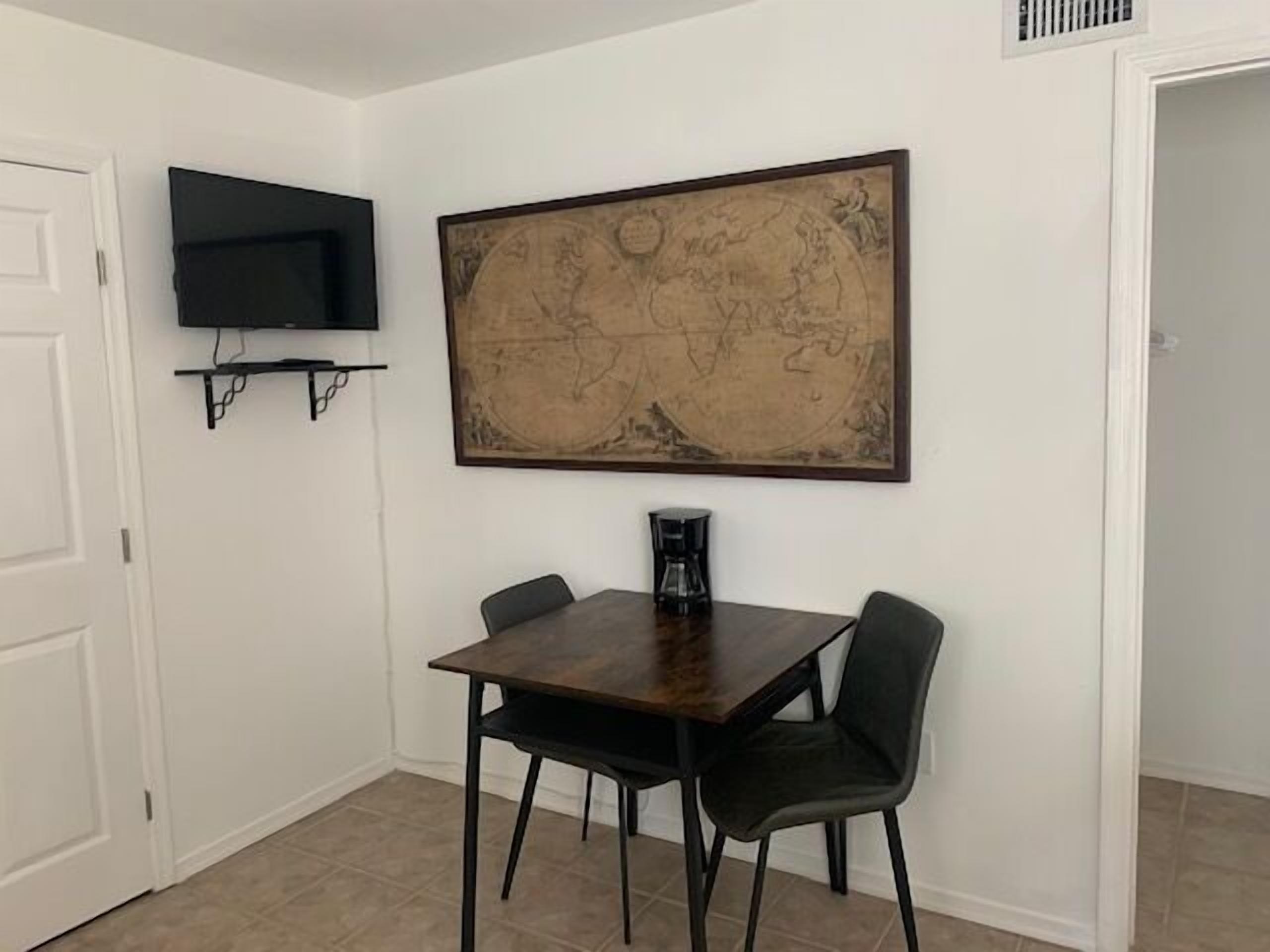 A dining area with a wooden table and two chairs, a large map on the wall, a television on a shelf, and a coffee maker on the table.
