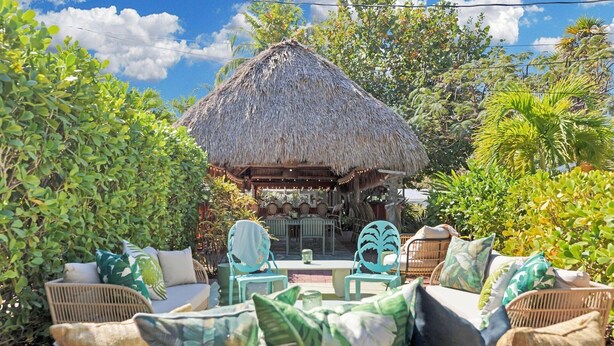 Un espacio al aire libre con mobiliario de mimbre y cojines, una estructura de techo de paja y abundante vegetación.