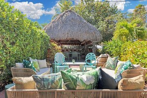 Un espacio al aire libre con mobiliario de mimbre y cojines, una estructura de techo de paja y abundante vegetación.