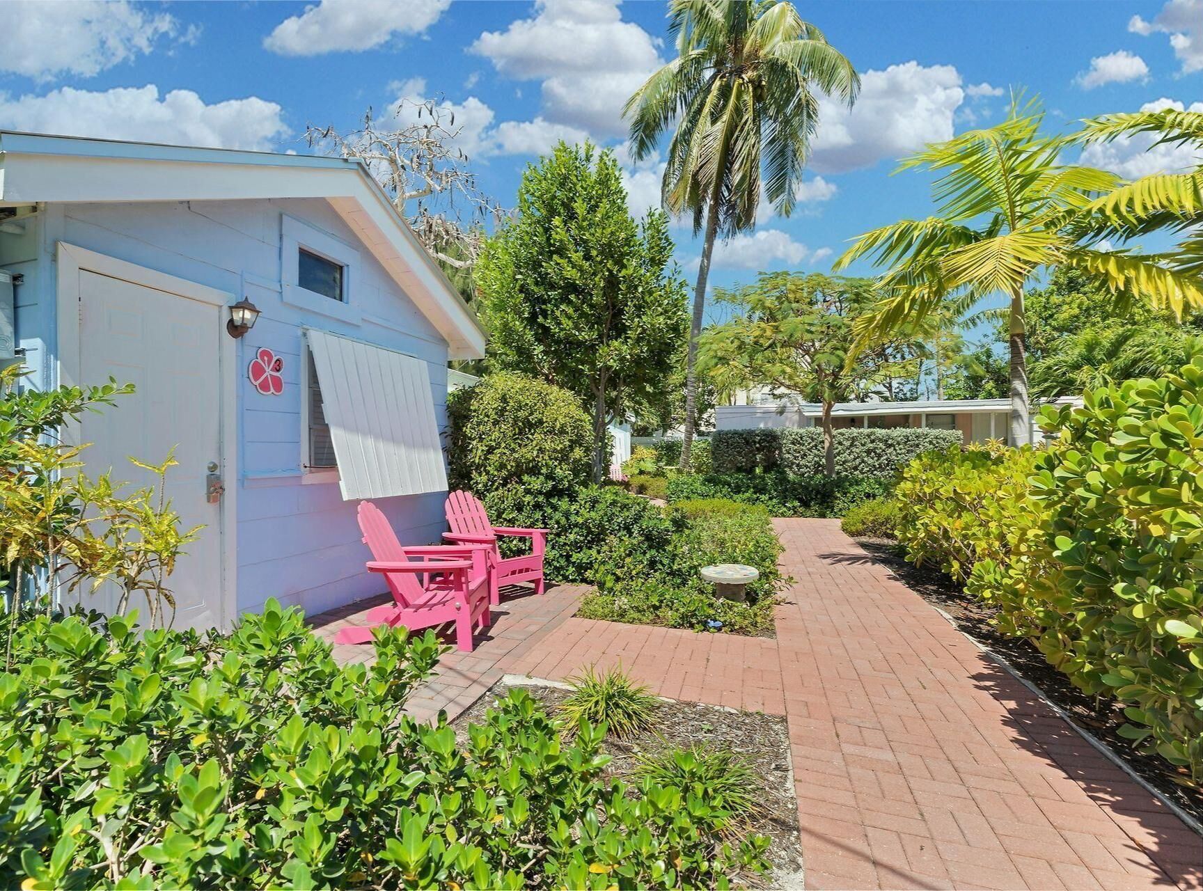 A small, light blue building with a door and windows, pink Adirondack chairs outside, and a brick pathway leading through a garden with various plants and a palm tree.