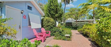 A small, light blue building with a door and windows, pink Adirondack chairs outside, and a brick pathway leading through a garden with various plants and a palm tree.