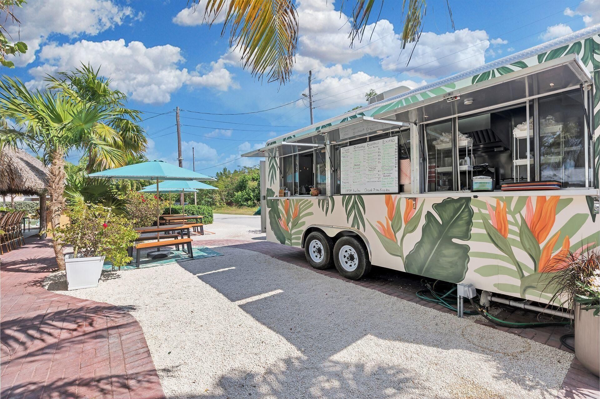 A food truck with a menu board, parked in a shaded outdoor area with palm trees and a thatched-roof structure.