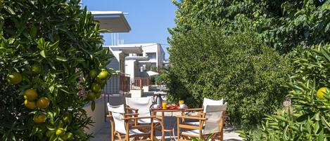 A patio with outdoor furniture and a table set for dining, surrounded by lush greenery and a modern building in the background.