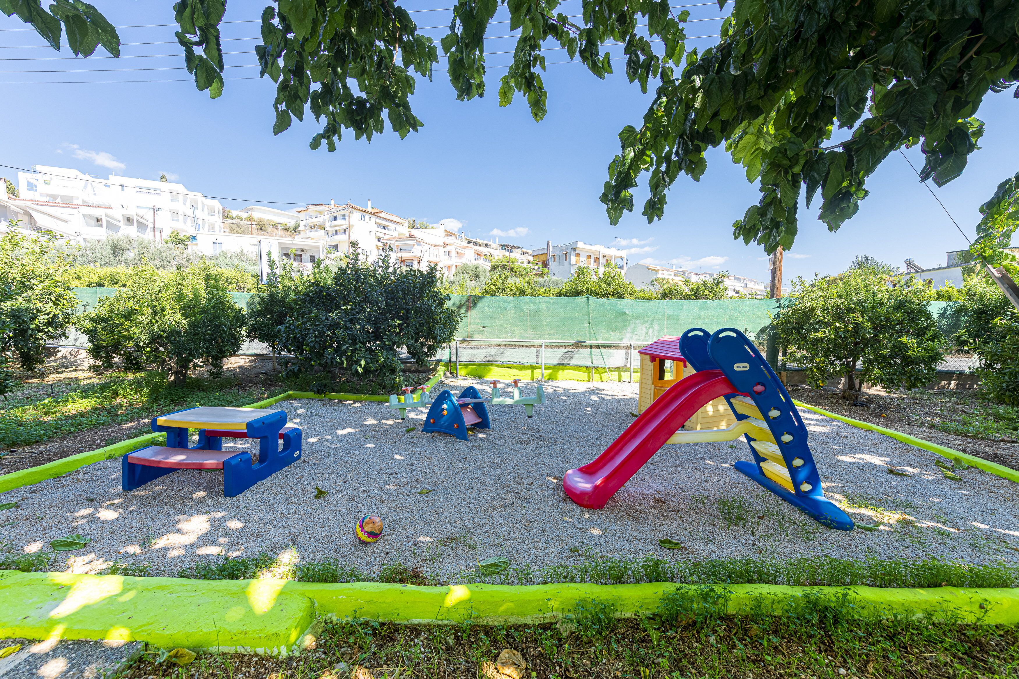 A playground with a slide and climbing structures, surrounded by a green fence and trees.