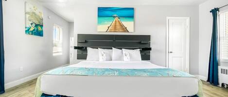 A hotel room with a large bed, a ceiling fan, a painting of a beach scene, and a window with curtains.