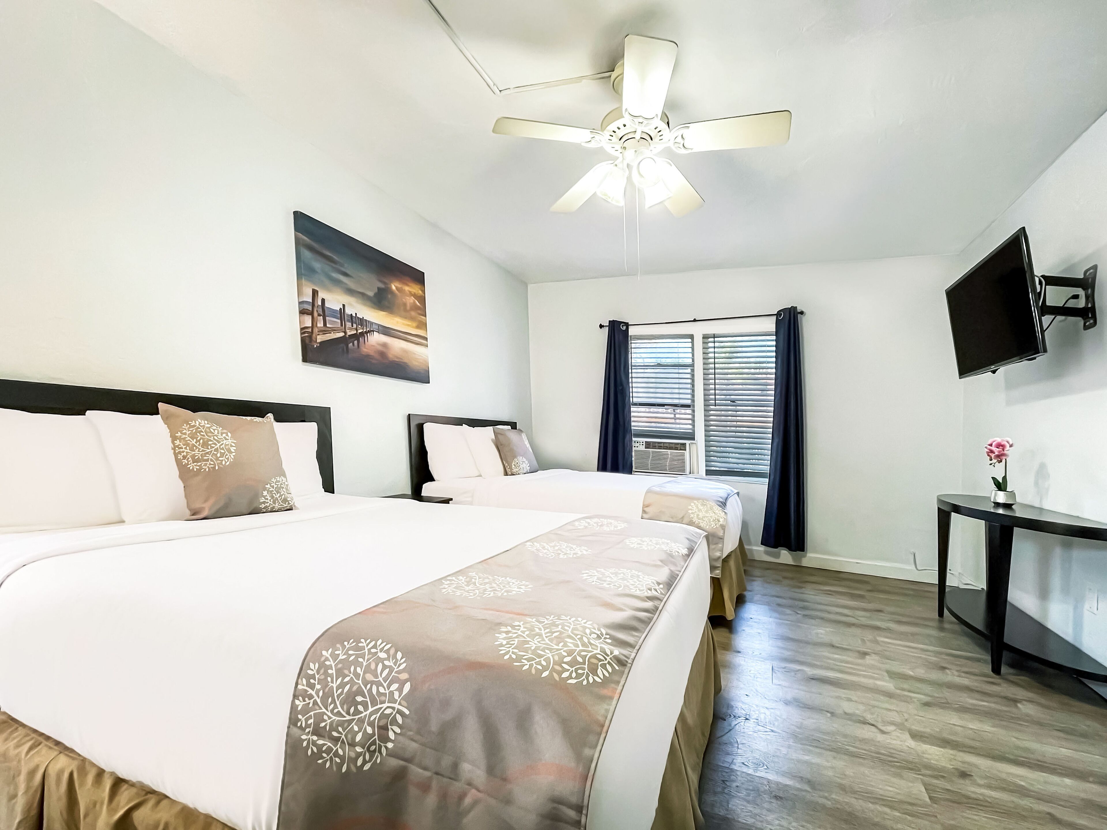 A hotel room with two beds, a ceiling fan, a painting, a TV, and a window with curtains.