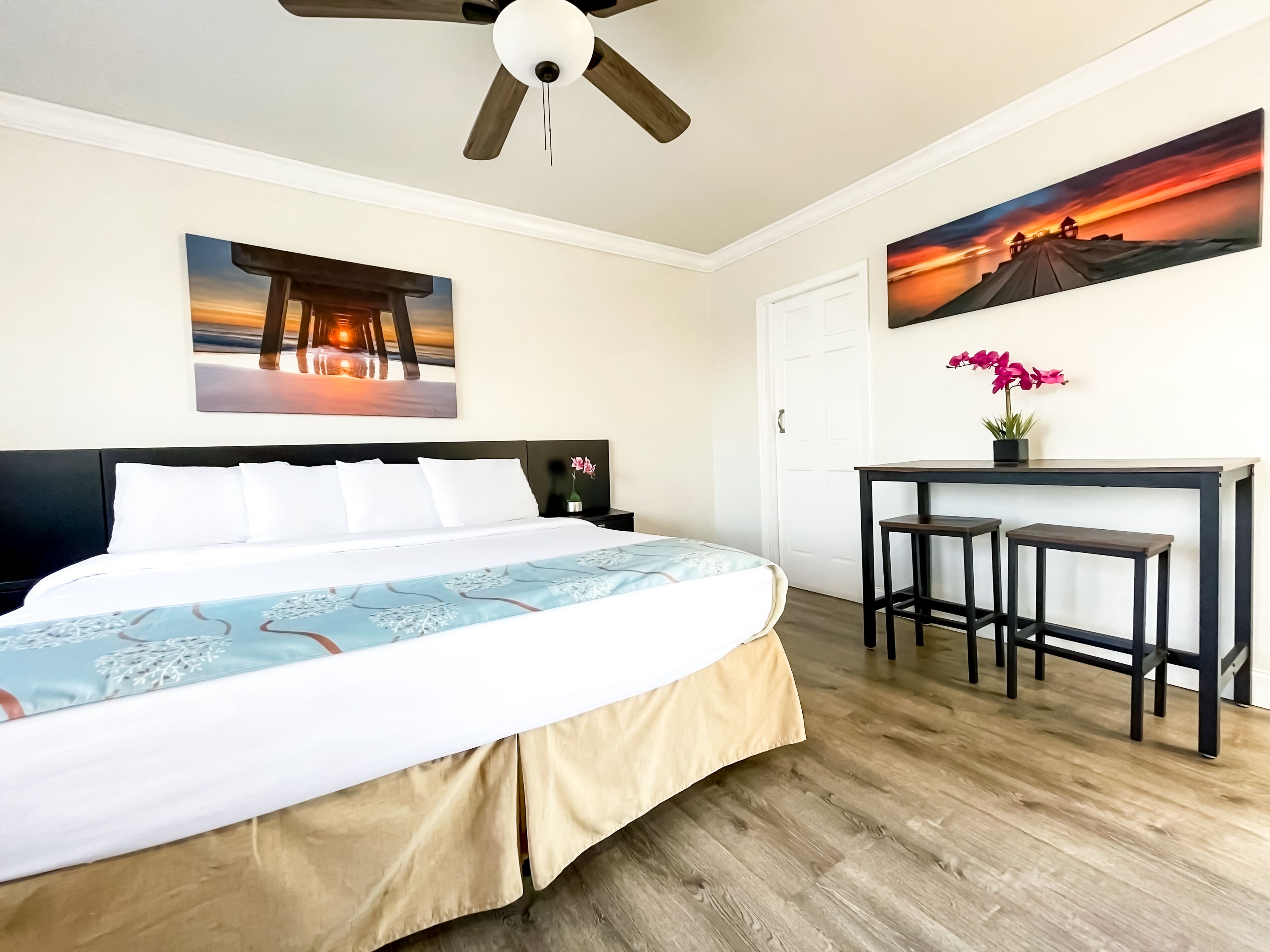 A hotel room with a bed, a desk with two stools, a ceiling fan, and two paintings on the walls.