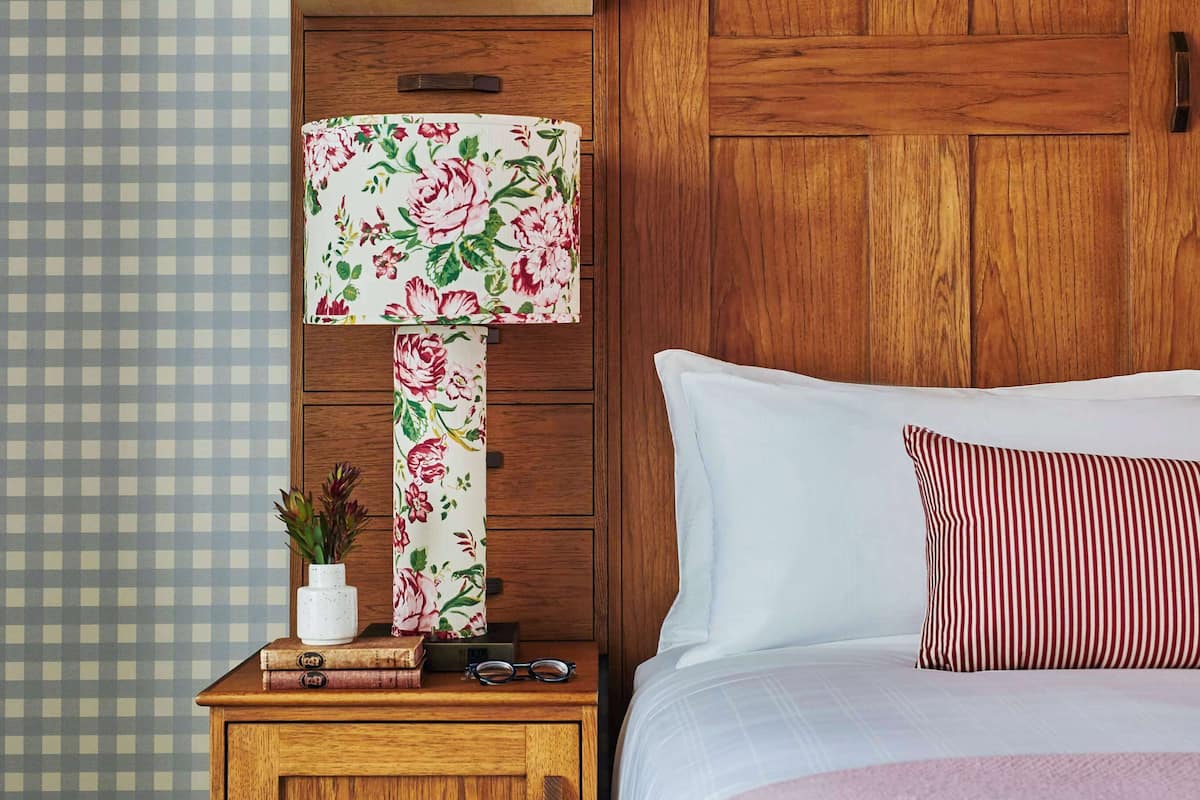 A bedroom with a wooden wardrobe, a bed with a striped pillow, a floral lamp, and a framed magazine on the wardrobe.
