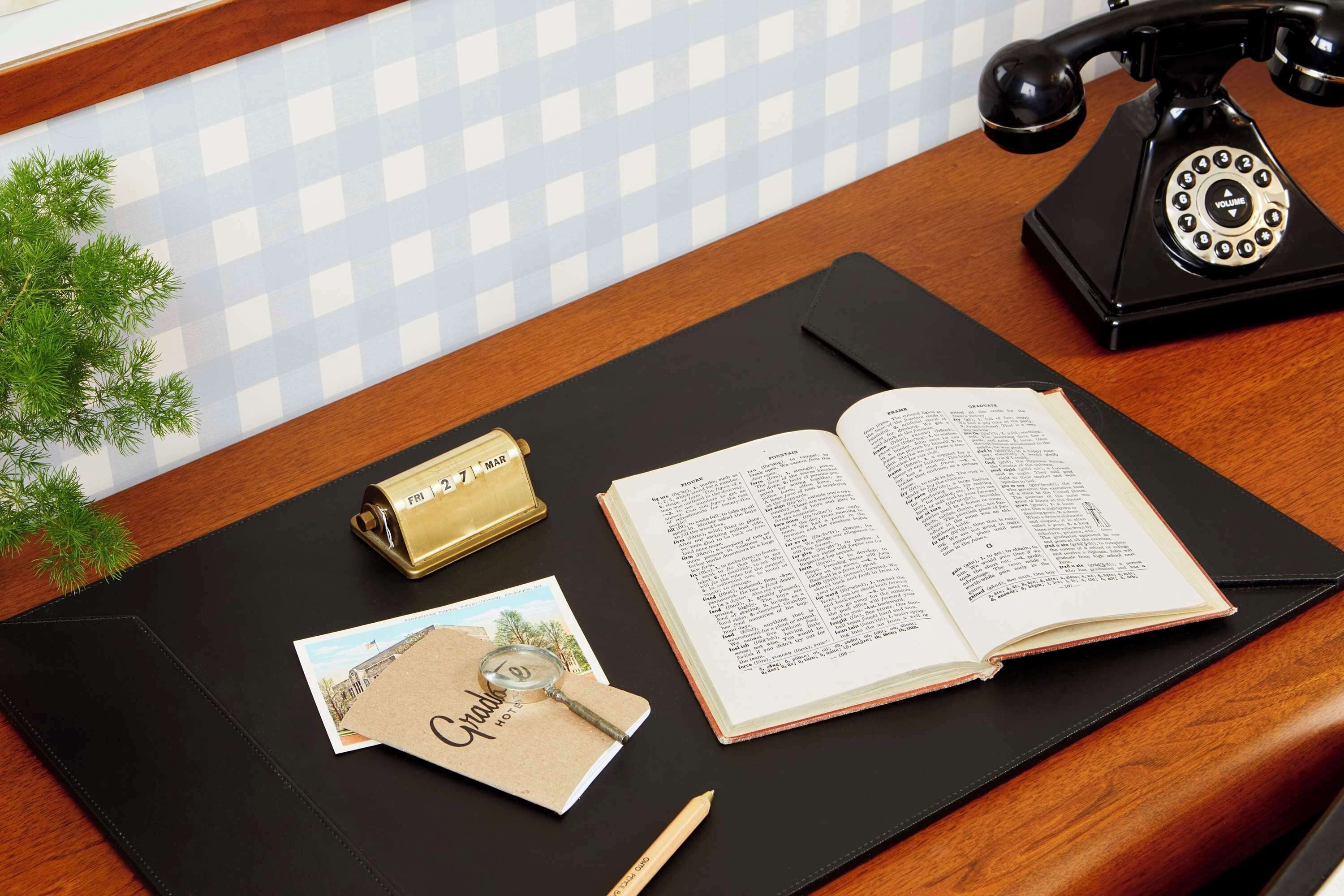 Un escritorio de madera con una libreta de cuero negro, una lupa, una postal, un calendario color dorado y un teléfono antiguo negro.