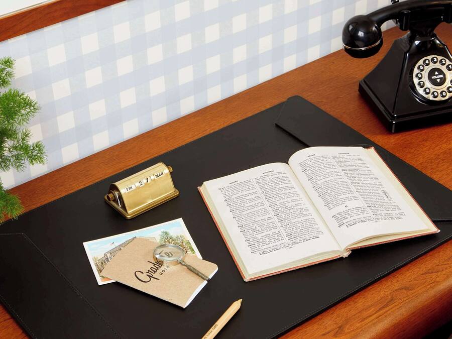 Un escritorio de madera con una libreta de cuero negro, una lupa, una postal, un calendario color dorado y un teléfono antiguo negro.