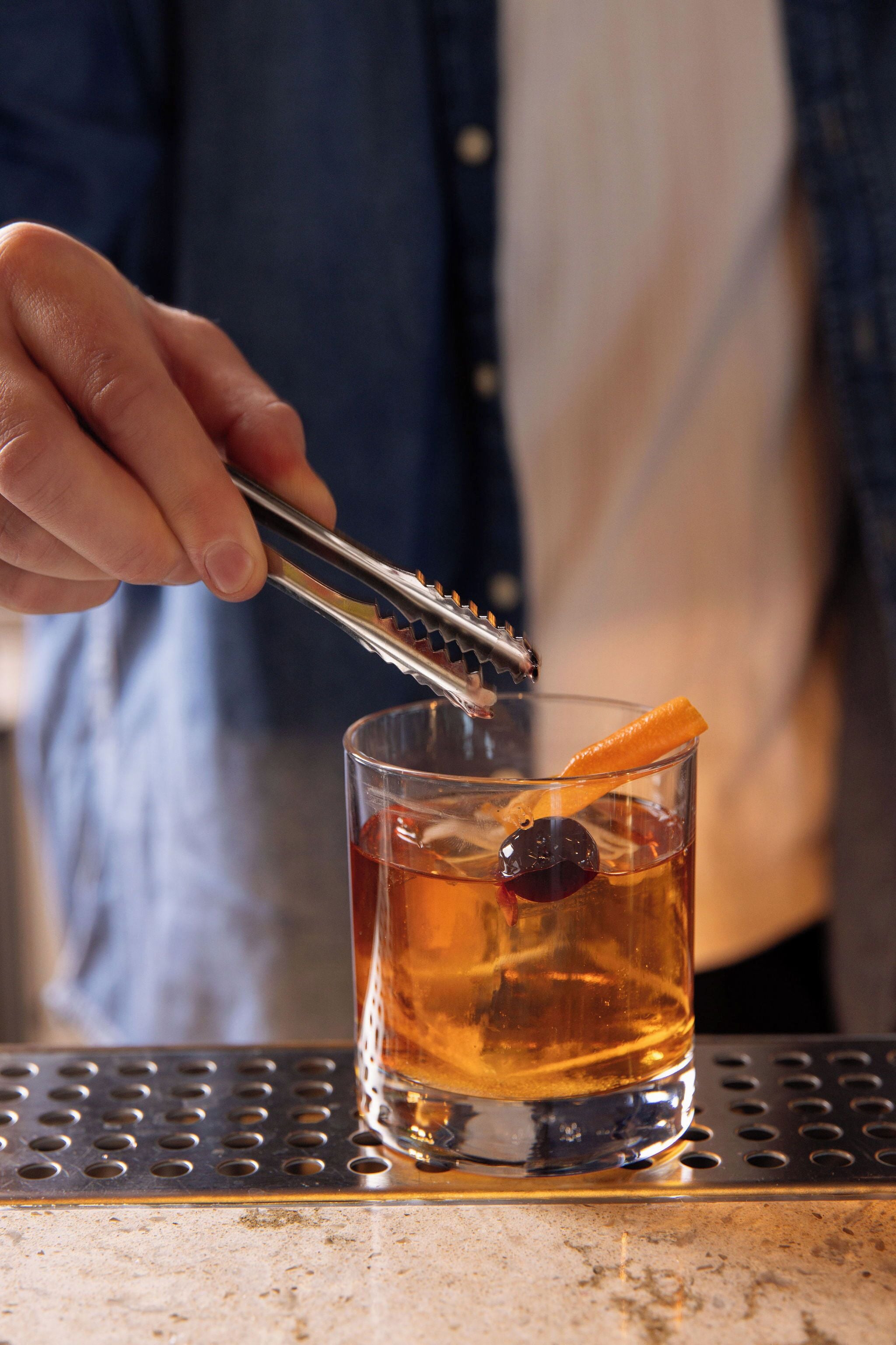 A person using tongs to add an orange slice to a glass of amber-colored liquid, likely a cocktail, on a metal bar counter.