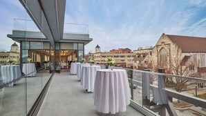 Um restaurante no terraço com mesas preparadas para refeições, com vista para uma paisagem urbana com edifícios históricos e um campanário.