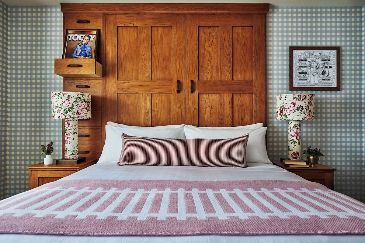A bedroom with a large wooden wardrobe, a bed with a pink and white striped bedspread, two bedside tables with floral lamps, and a framed picture on the wall.