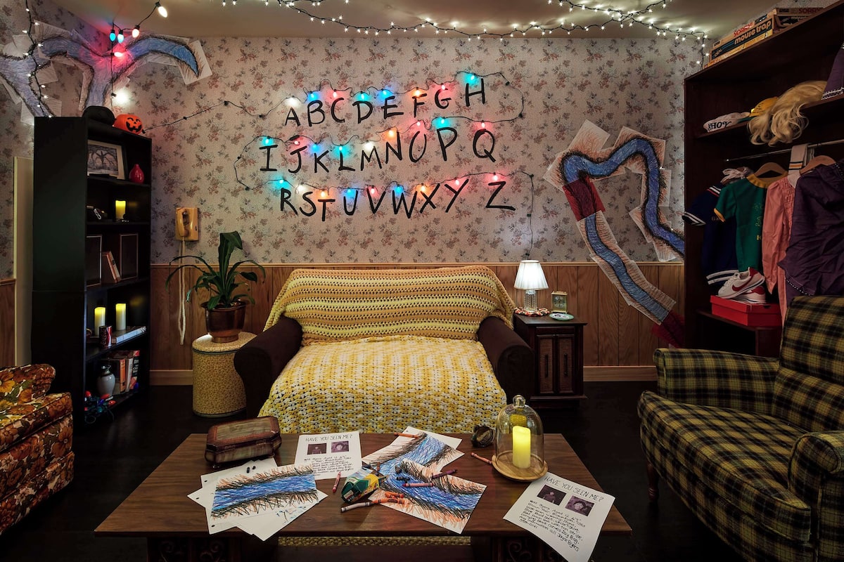 A cozy room with a bed, a table with papers, a bookshelf, and a wall with illuminated letters.