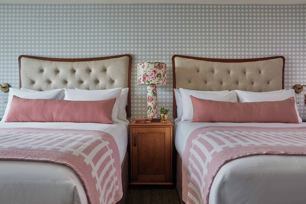 A hotel room with two beds, each with a tufted headboard, white bedding, and red and white striped pillows. There is a wooden nightstand with a lamp between the beds.