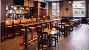 Um restaurante com mesas e cadeiras de madeira, área de bar com garrafas e plantas, e janelas amplas com cortinas.